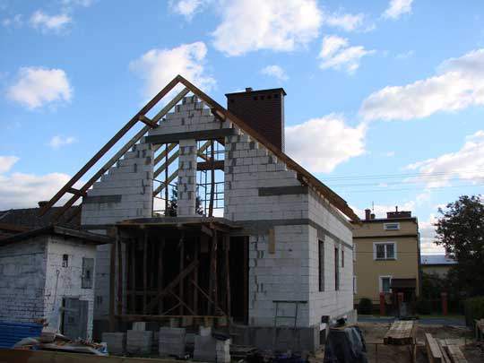 Surowy stan budowy domu jednorodzinnego z białych bloczków, widoczne drewniane elementy konstrukcji dachu i komin z cegły, na tle błękitnego nieba z obłokami.